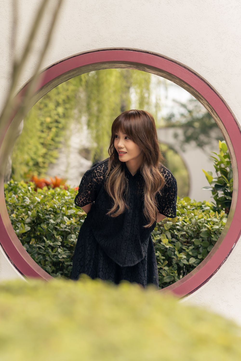 A person in a black lace dress poses through a circular garden window frame among green foliage.