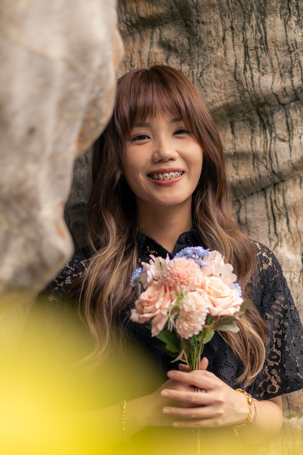 A person in black holds a bouquet of pink flowers while standing by a tree.