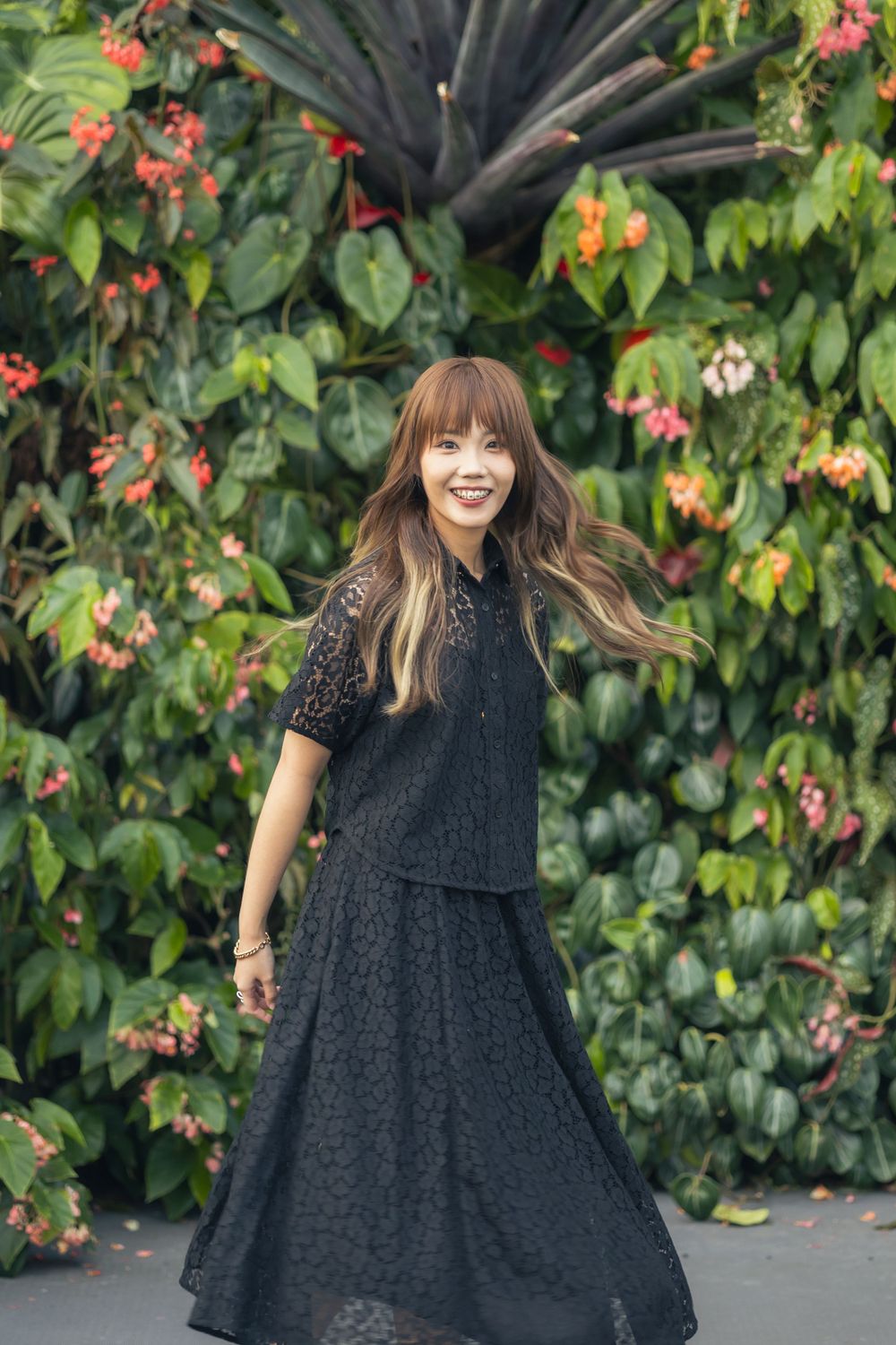 A person in a black lace ensemble twirls joyfully against a backdrop of tropical plants with red flowers.