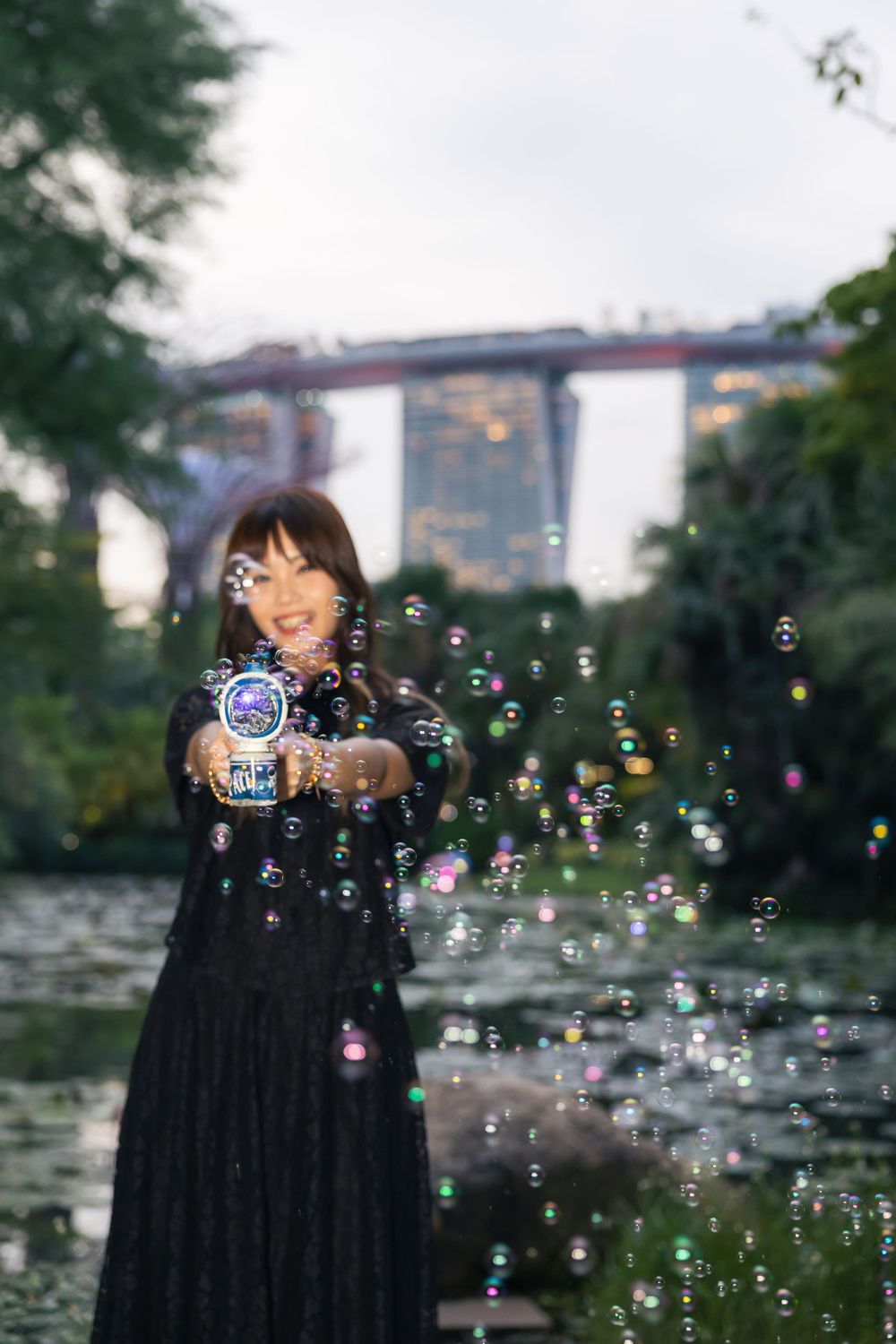 A person in a black dress creates bubbles at Gardens by the Bay with Marina Bay Sands visible in background.