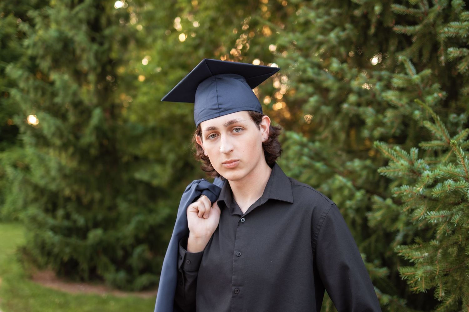 Graduate wearing blue mortarboard cap and gown stands thoughtfully against green foliage background outdoors.