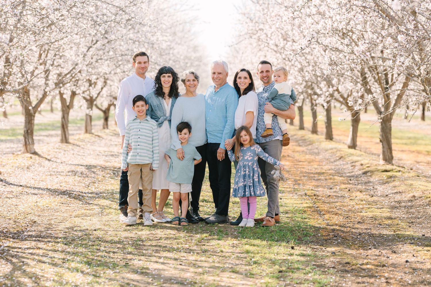Extended family gathering for photos among blooming almond trees in orchard setting.