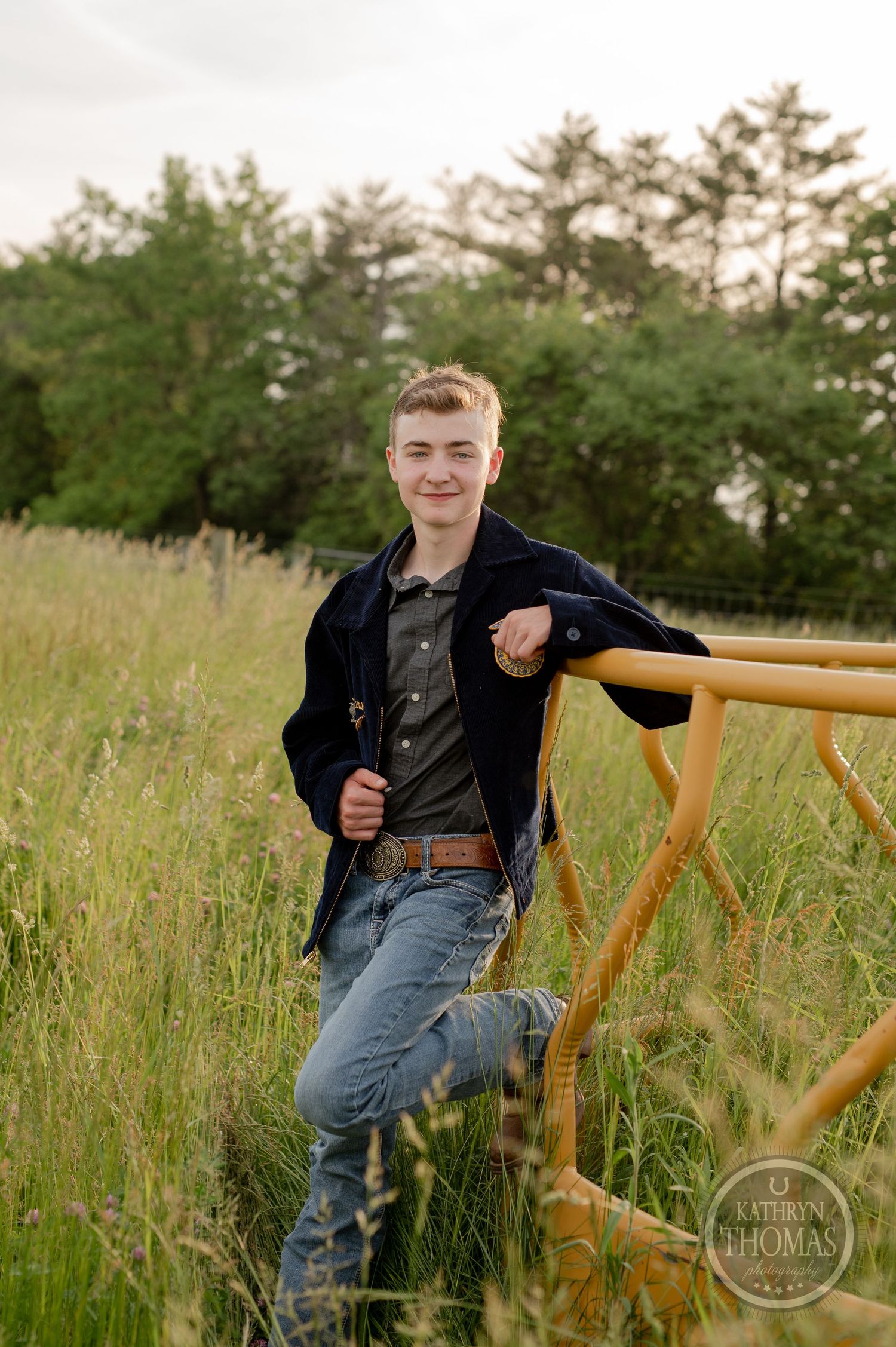 Student in navy FFA jacket poses by yellow railing in scenic grassland during golden hour.