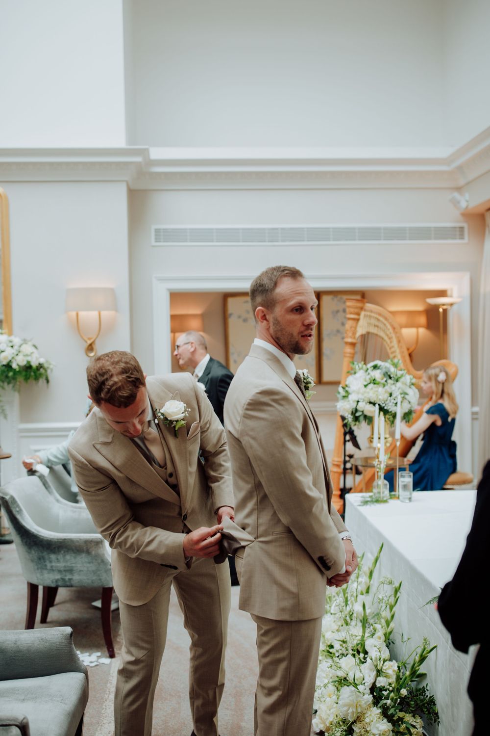 Two groomsmen in tan suits standing in an elegant room decorated with white floral arrangements at a formal event.