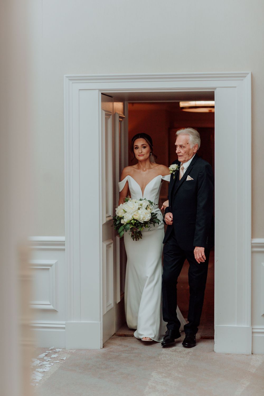A bride in an elegant white off-shoulder dress holds a bouquet while walking through a doorway with an older man in a suit.