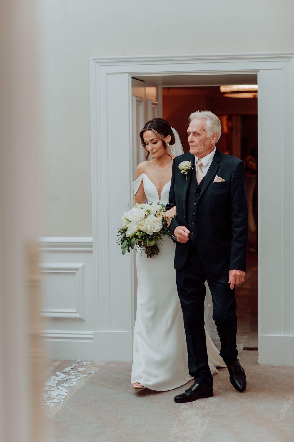 A father escorting a bride in an elegant white gown while holding a beautiful white floral bouquet through a doorway.