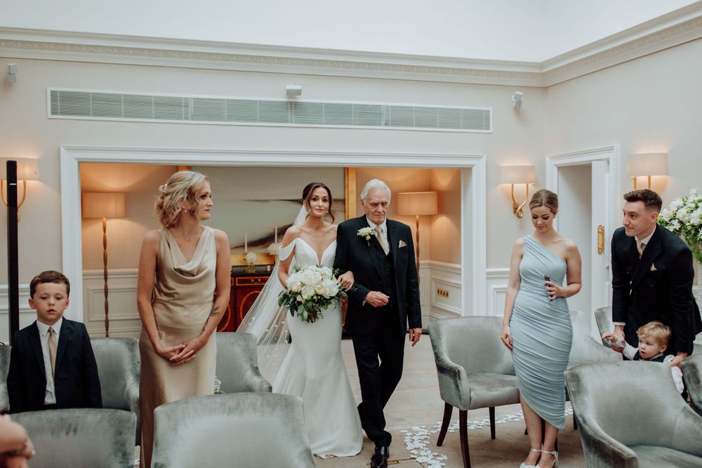 A wedding party stands together in an elegant white hotel room with gray upholstered chairs.