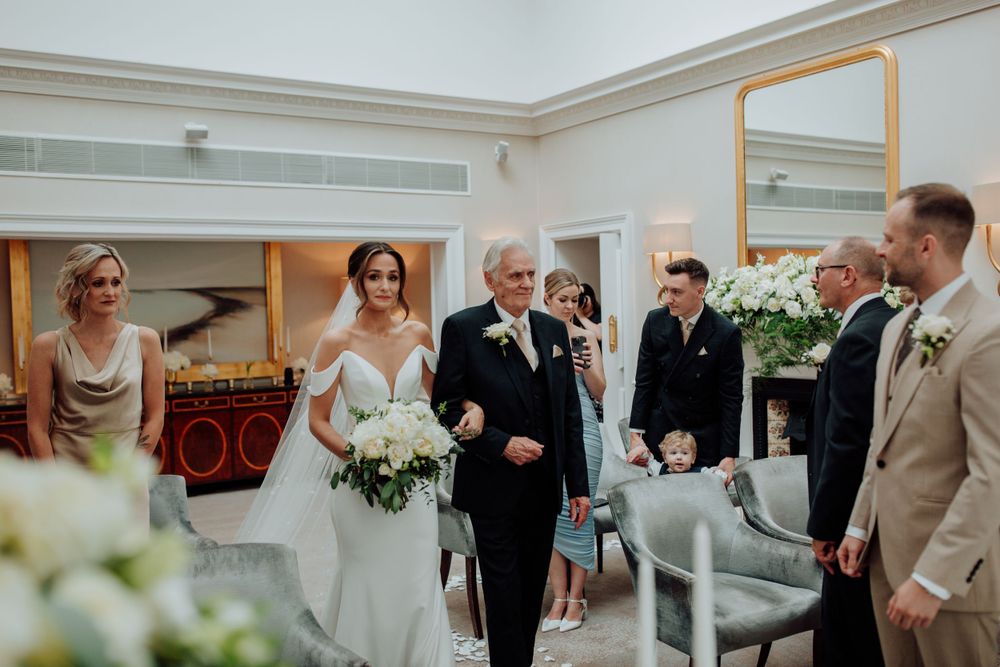 Elegant wedding ceremony takes place in a bright indoor venue decorated with white flowers and soft furnishings.
