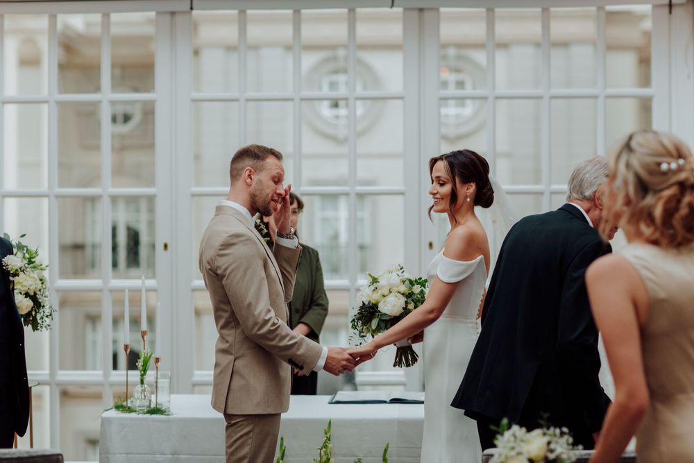 A couple exchanges vows during their wedding ceremony inside an elegant conservatory with white greenhouse windows.