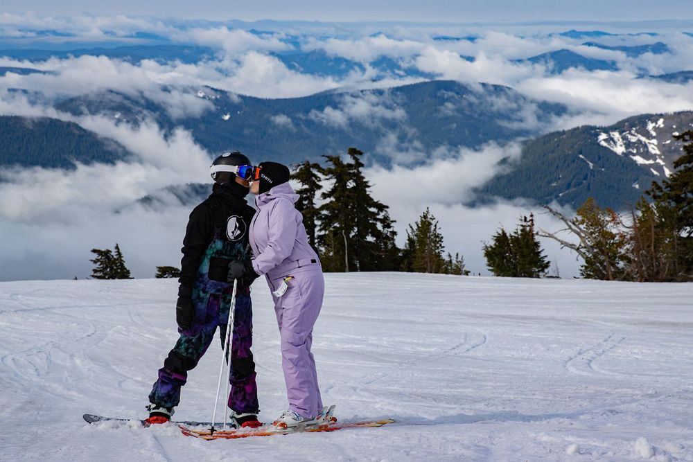Two skiers stand together on a snowy mountain slope above the clouds on a sunny day.