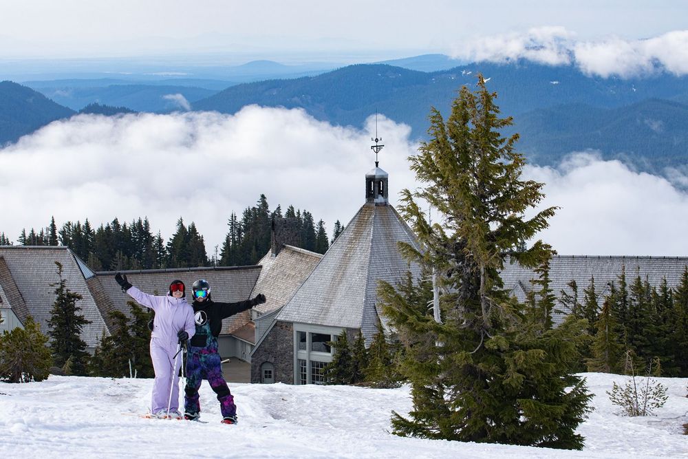 Two snowboarders pose on a snowy Mt.Hood slope with Historic Timberline Lodge and cloud-covered peaks in the background.