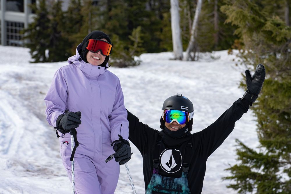 Two skiers in colorful snow gear smile and pose on a snowy mountain slope surrounded by pine trees.