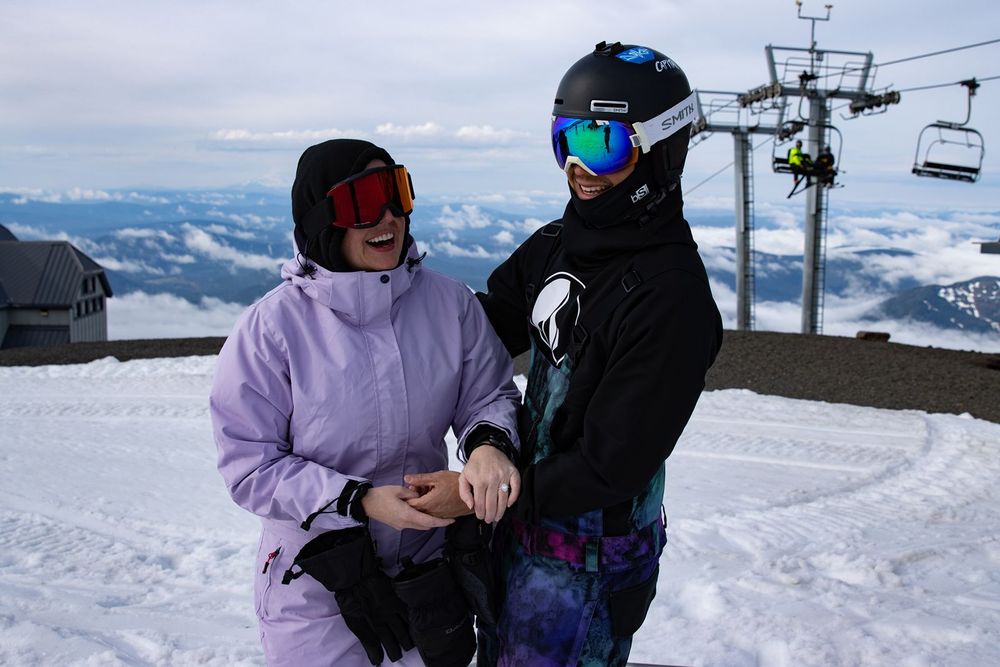 Two snowboarders in colorful winter gear laugh together on a snowy mountain slope with ski lifts in the background.