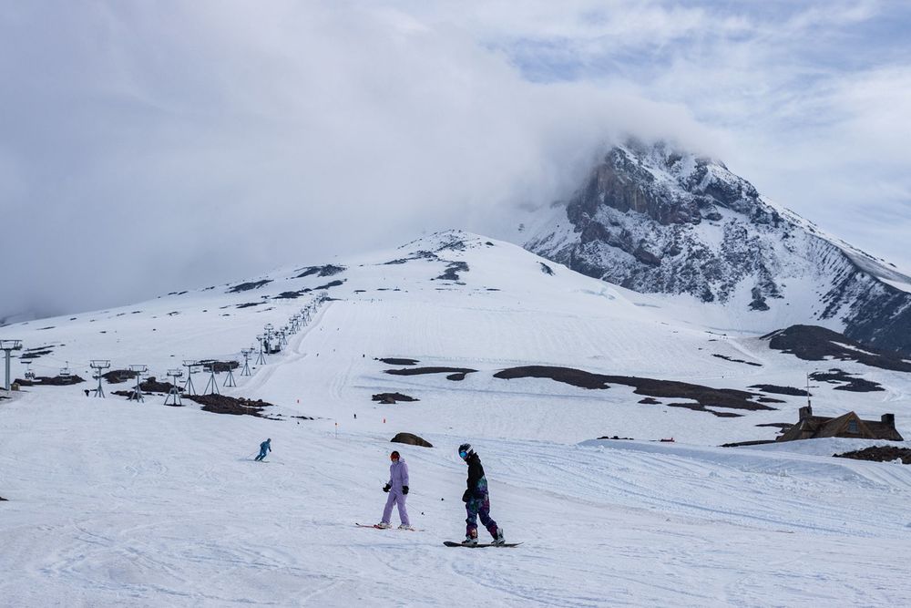 Skiers on a snowy slope with a dramatic snow-capped Mt.Hood peak emerging through clouds in the background.