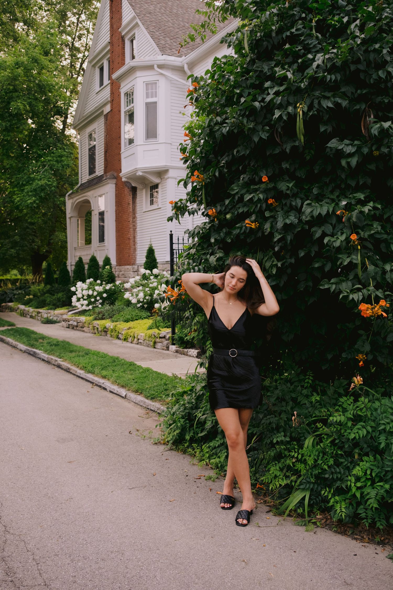 Stylish black dress pose in front of a Victorian house with landscaped gardens.