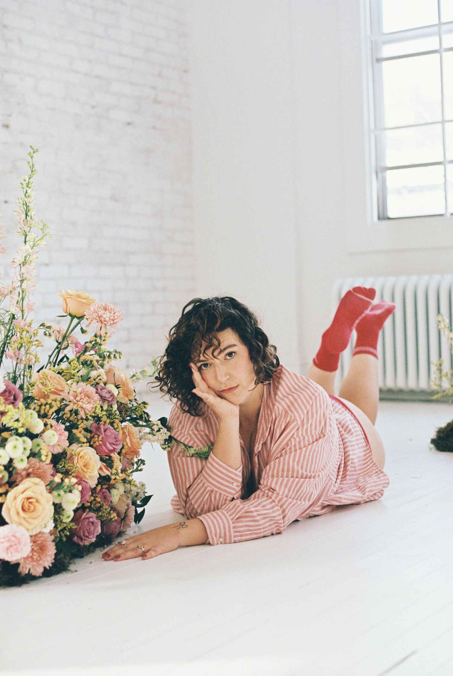 Boudoir photo with person lying next to floral arrangement in pink striped shirt.