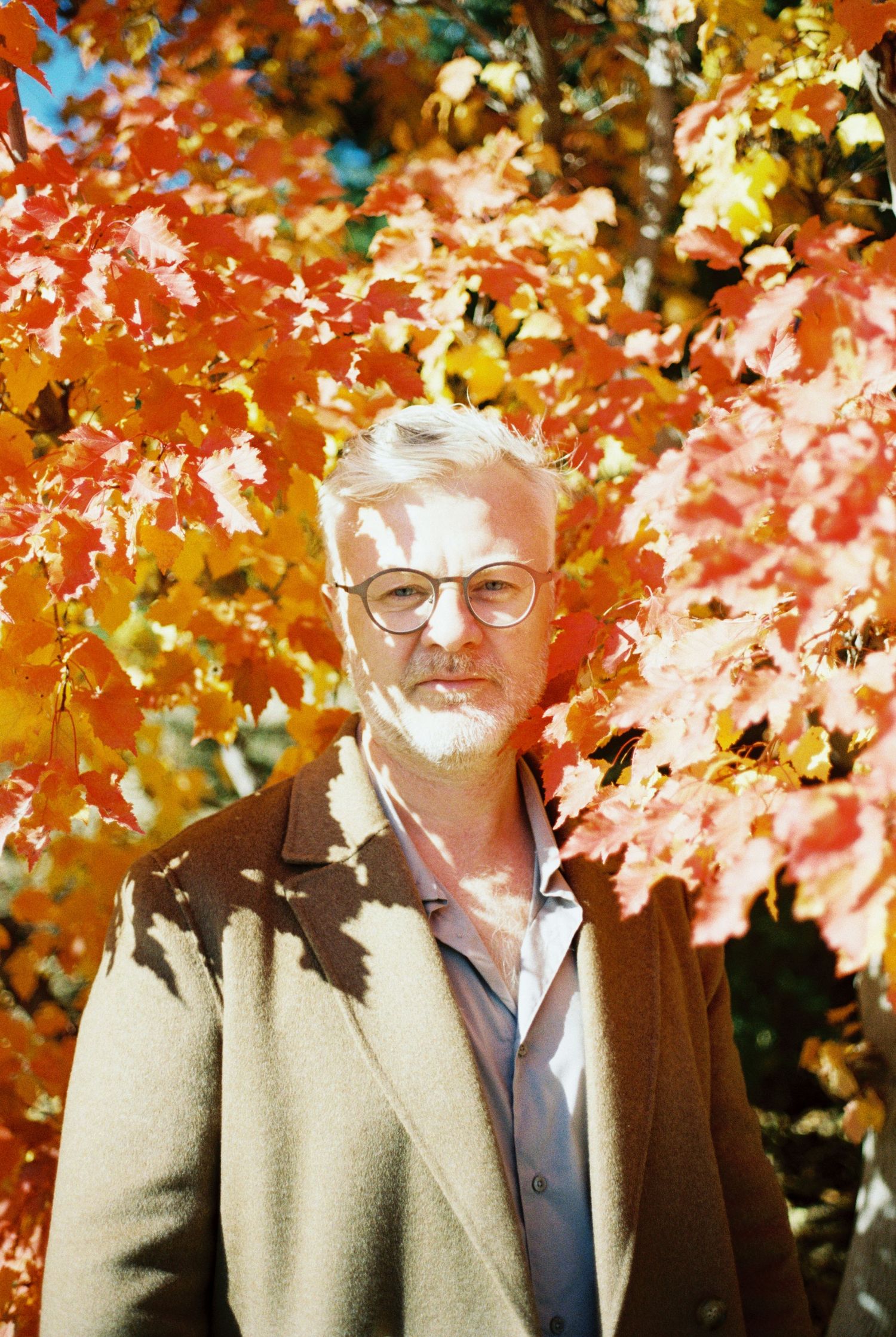 Portrait against vibrant autumn foliage wearing beige coat and glasses.