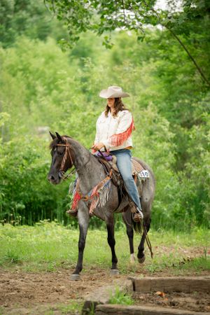 Equestrian riding gray horse with decorative fringe tack through lush green forest trail.