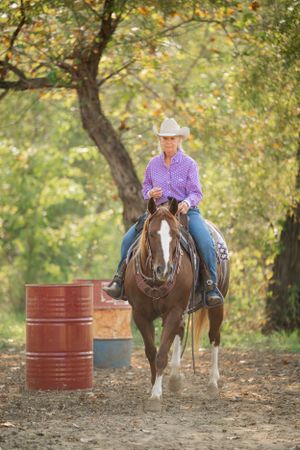 Horse and rider navigating barrel racing course on dirt arena with autumn trees in background.