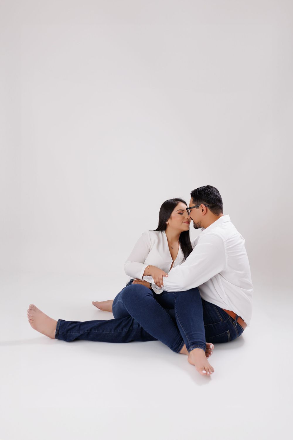 Couple in white shirts and jeans share intimate moment on white background.