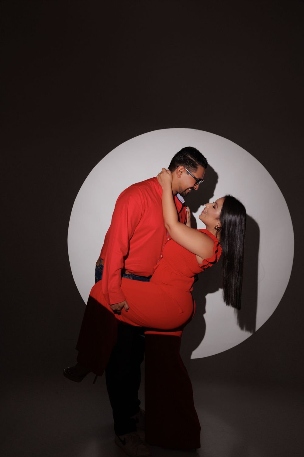Dramatic silhouetted couple in red outfits pose romantically against circular light.