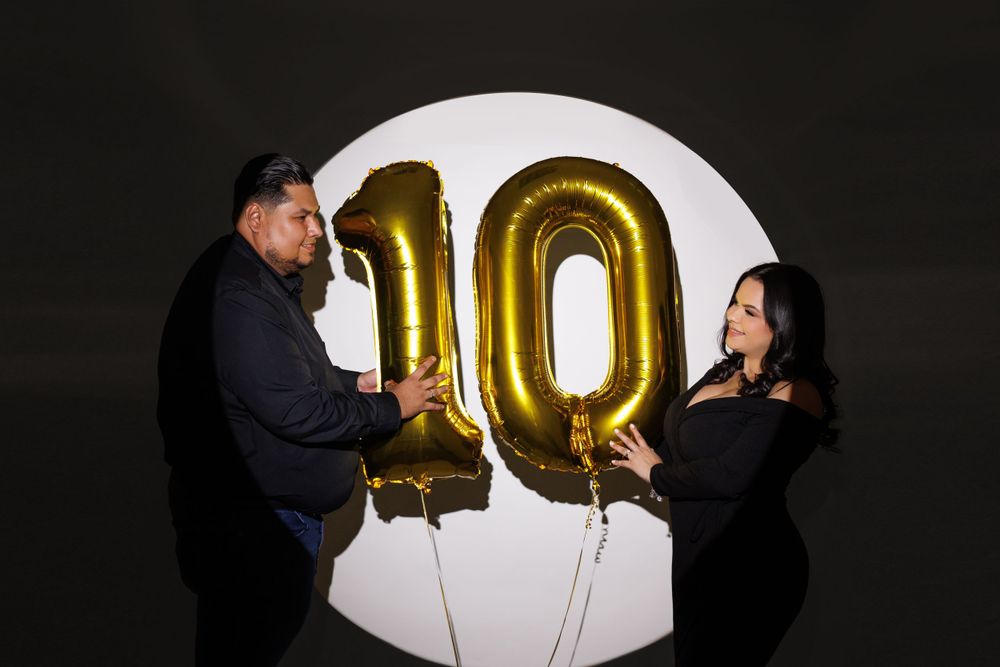 Estamos especializados en fotografía de bebés y famA couple holds large gold number 10 balloons against a dark backdrop with circular light.