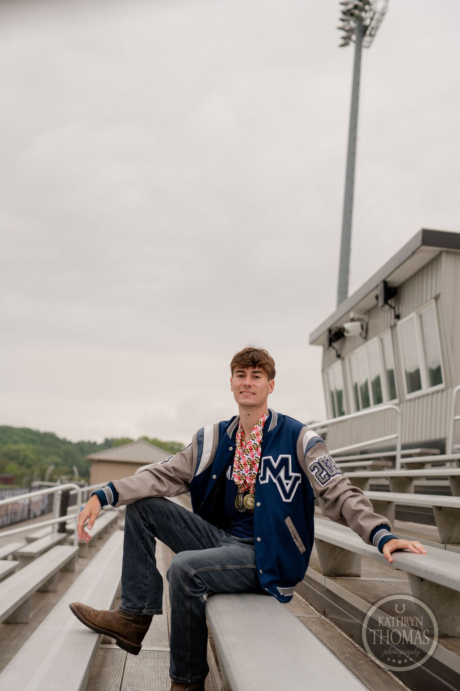 Student in varsity letterman jacket relaxes on stadium bleachers on an overcast day.