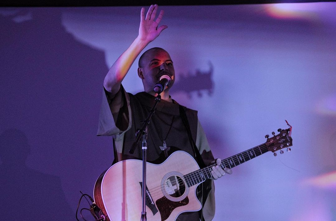 Catholic Priest performs on stage with acoustic guitar under purple lighting while raising hand to audience.