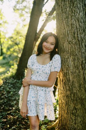 A person in a floral white dress poses against a large tree trunk in a sunlit forest setting.