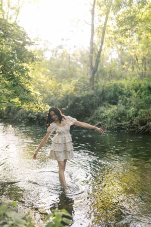 A person in a white dress stands playfully in a shallow stream surrounded by lush greenery.