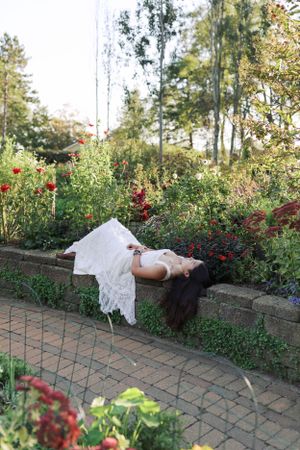 A figure in a white dress lies artistically on a brick retaining wall surrounded by blooming garden flowers.