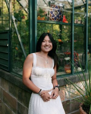 A person in a white summer dress poses by a glass greenhouse with decorative hanging ornaments.