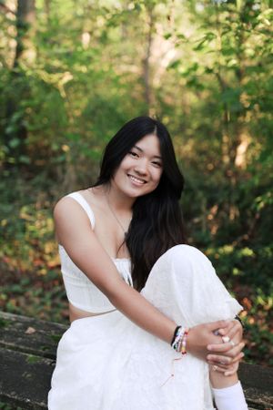 Person in white dress sitting casually on wooden surface in outdoor forest setting.
