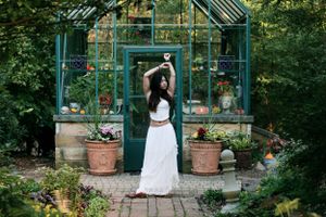 Figure in white outfit poses in front of green greenhouse with potted plants.