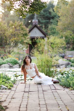 Person in white dress poses on brick path near decorative garden building and pond.