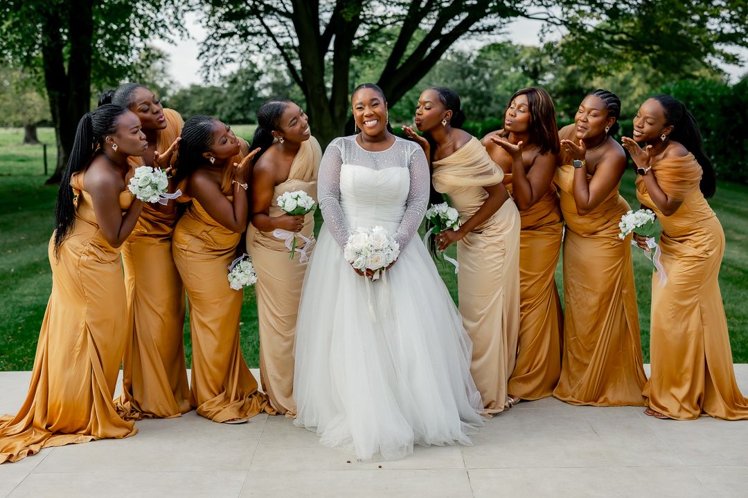 Wedding party poses outdoors with bride in white gown surrounded by bridesmaids in golden dresses.