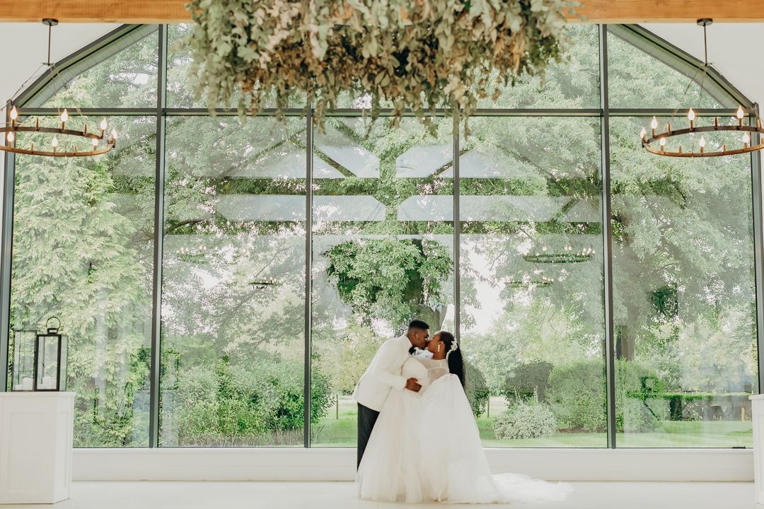Wedding couple dances in glass-walled venue decorated with hanging greenery and florals.