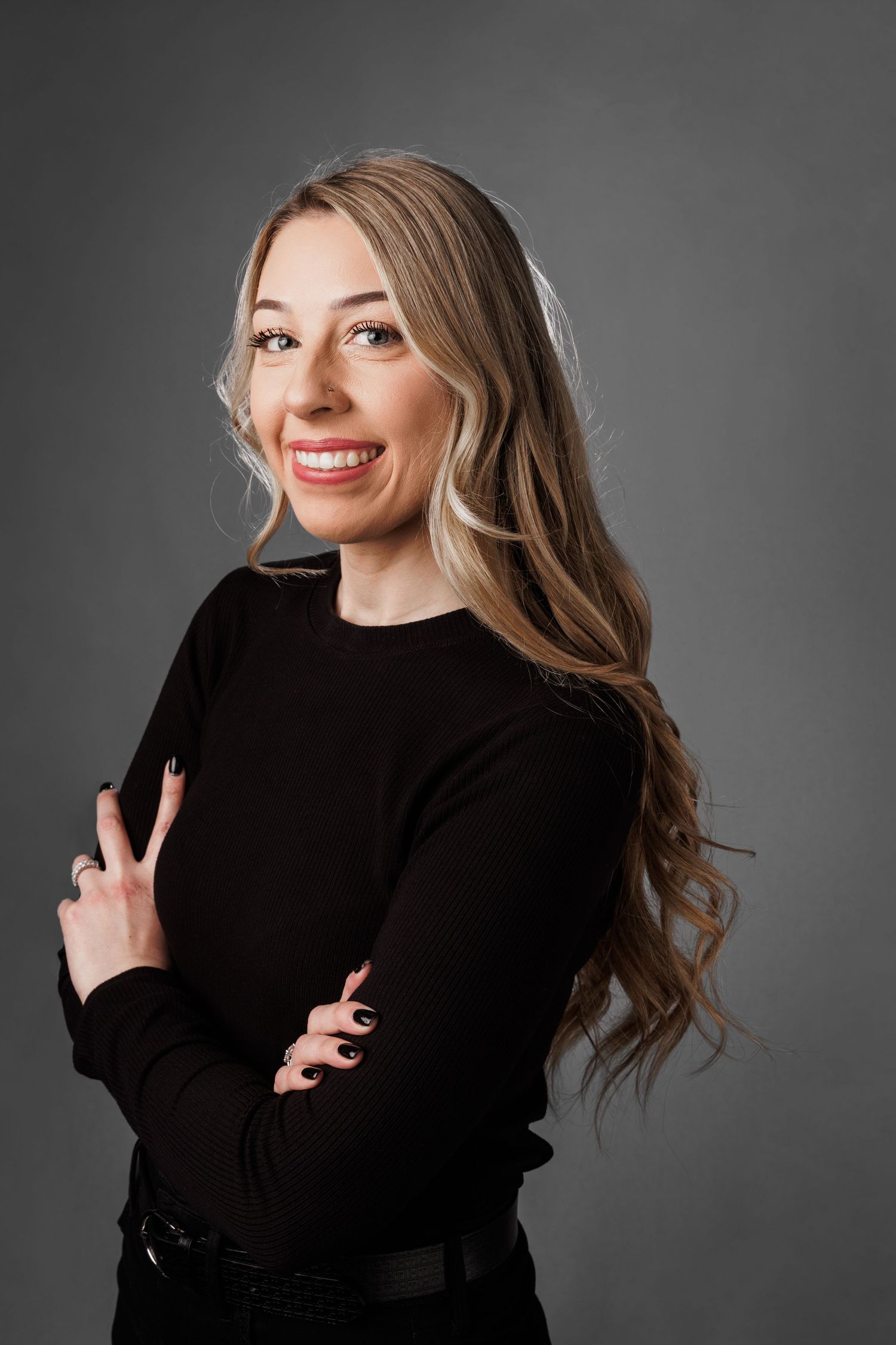 Smiling blonde woman in black long-sleeve top with arms crossed, posing against a gray studio background.