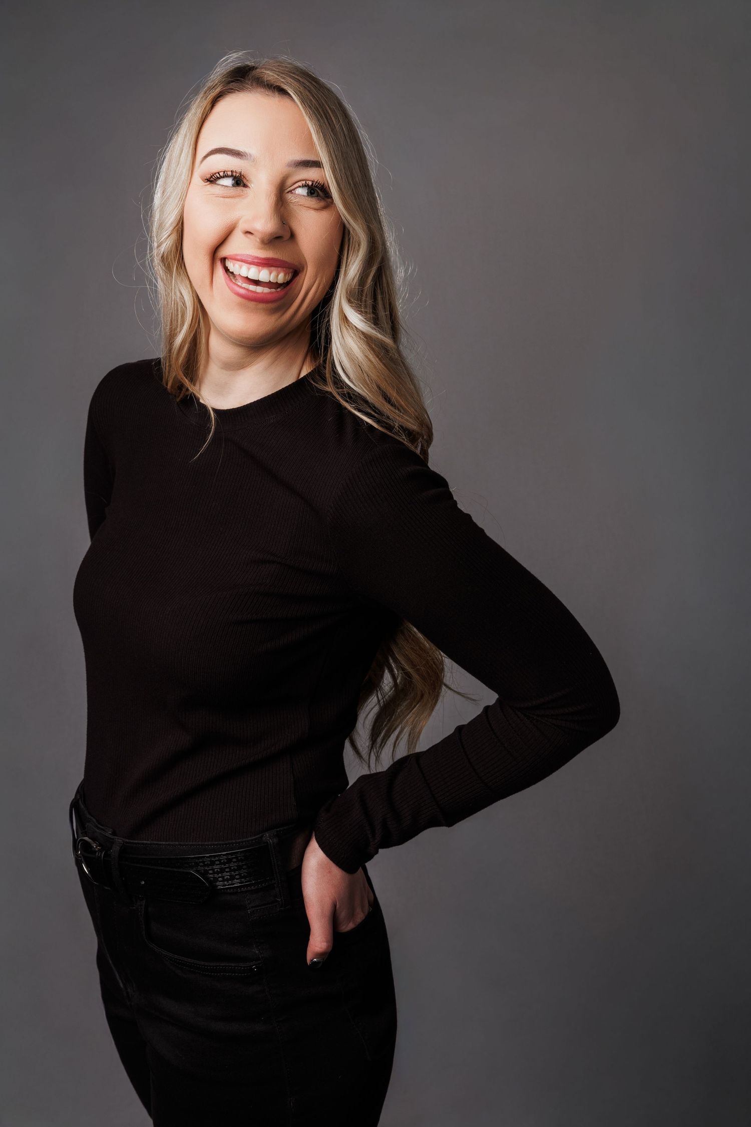 Smiling blonde woman in black long-sleeve top and jeans posing with hand on hip against gray background.