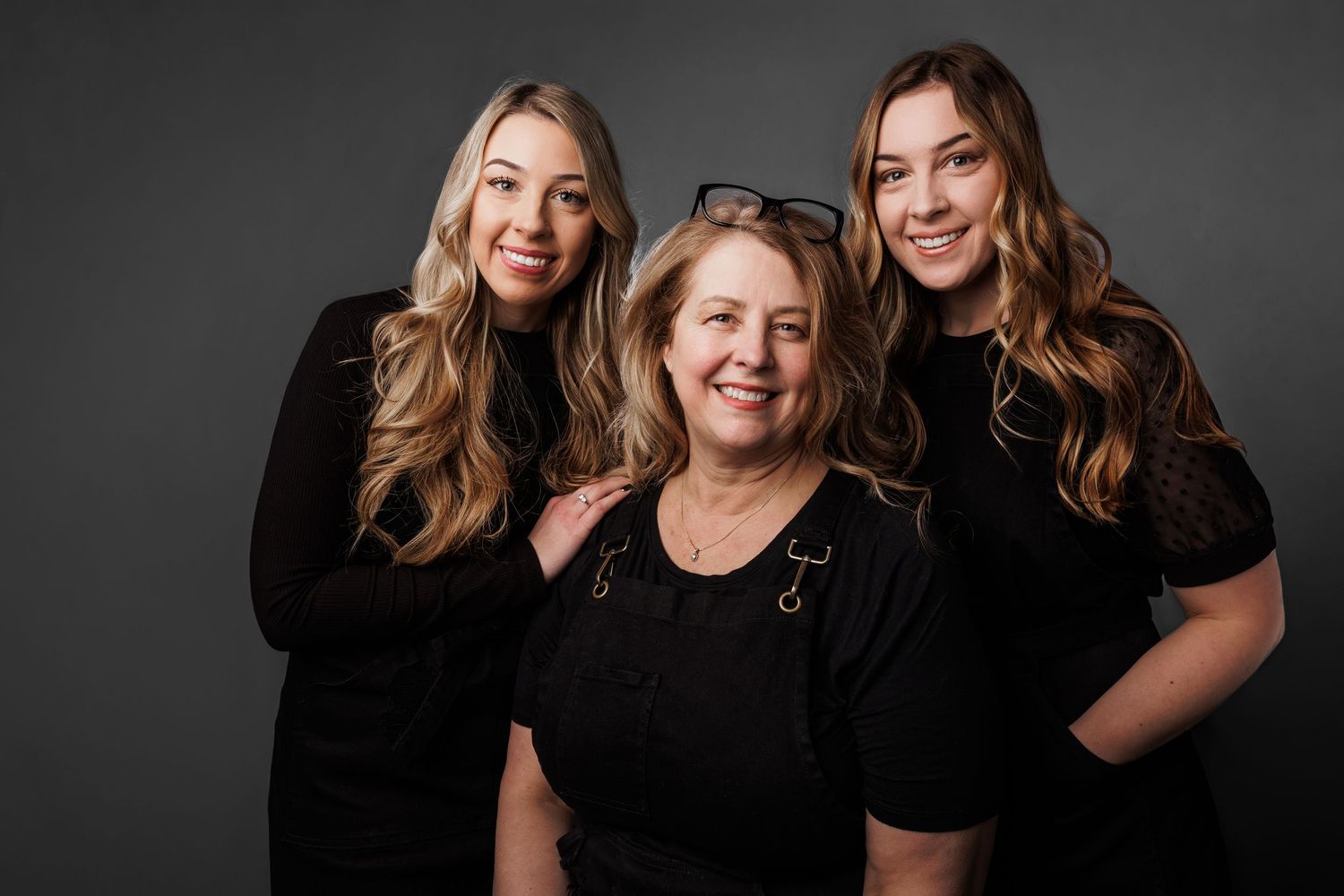 Three women in black outfits pose together for a professional portrait against a dark gray background.