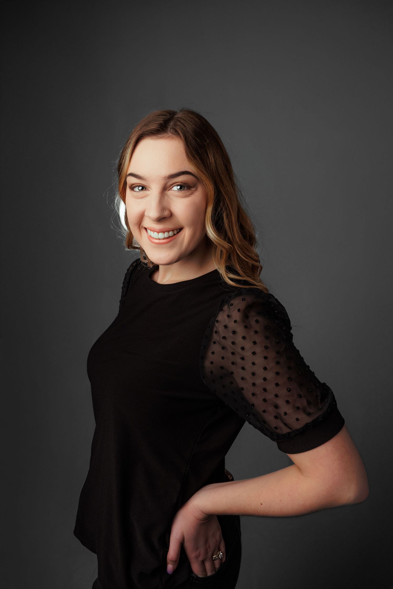 Smiling young woman in black polka dot sheer sleeve top posing confidently against a gray studio background.