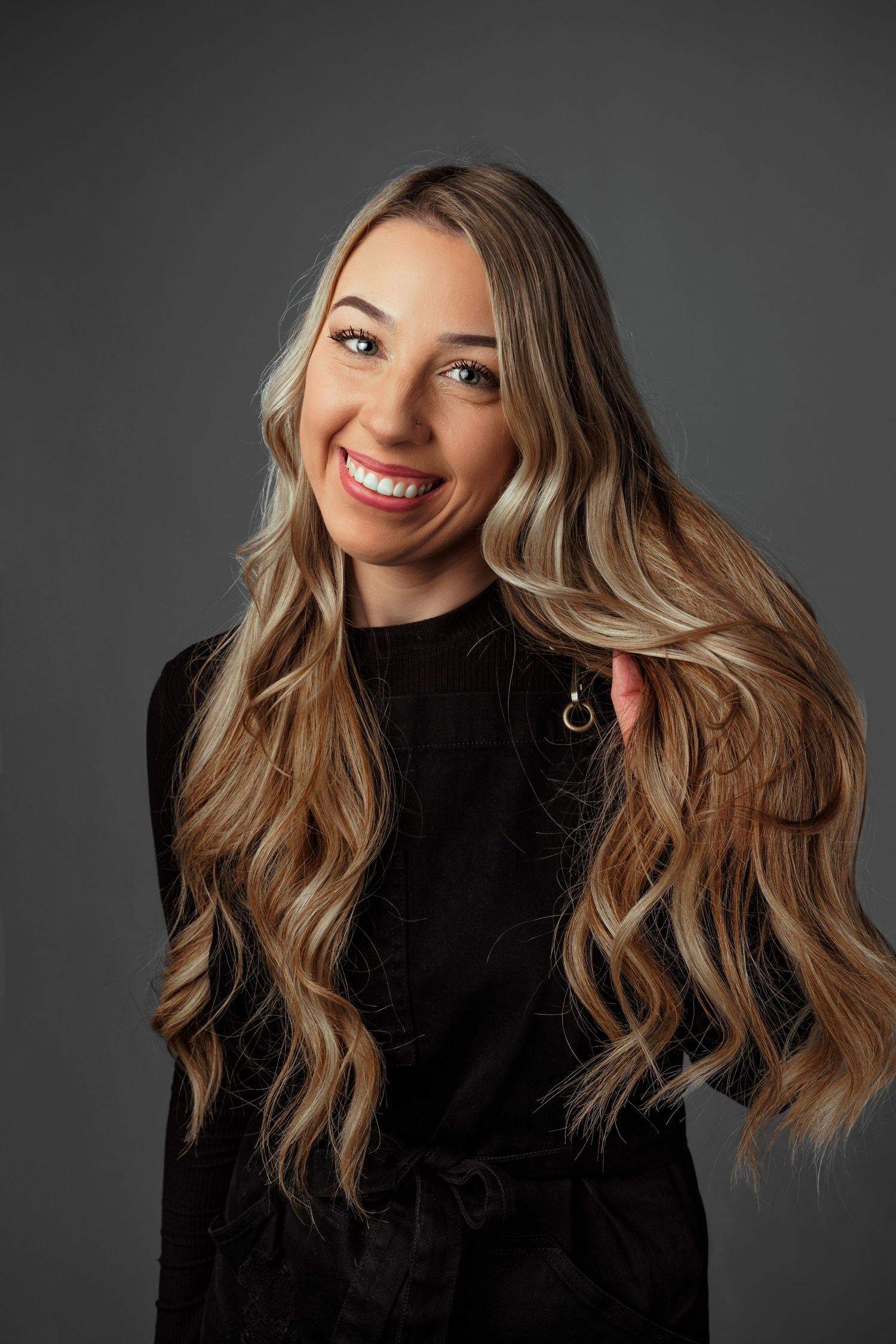 Smiling young woman with long wavy blonde highlighted hair wearing a black top, posing against a gray background.