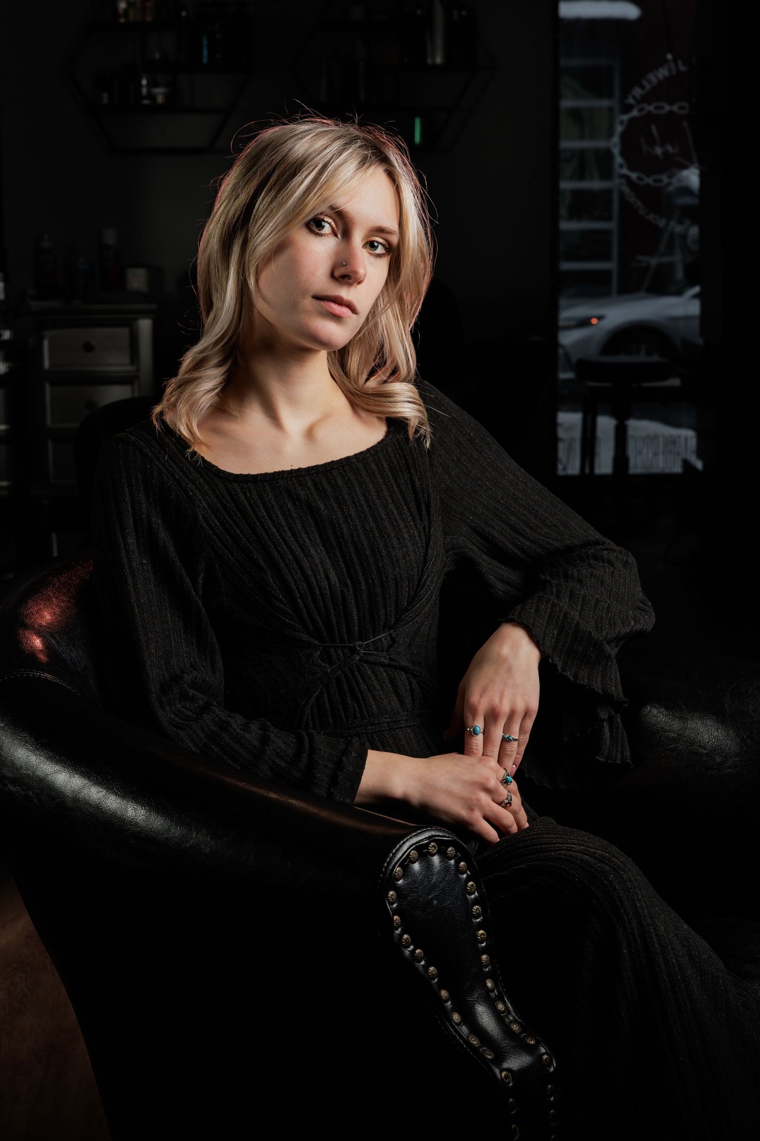 Blonde woman in black dress seated in dark leather chair, posing in moody dimly lit room.
