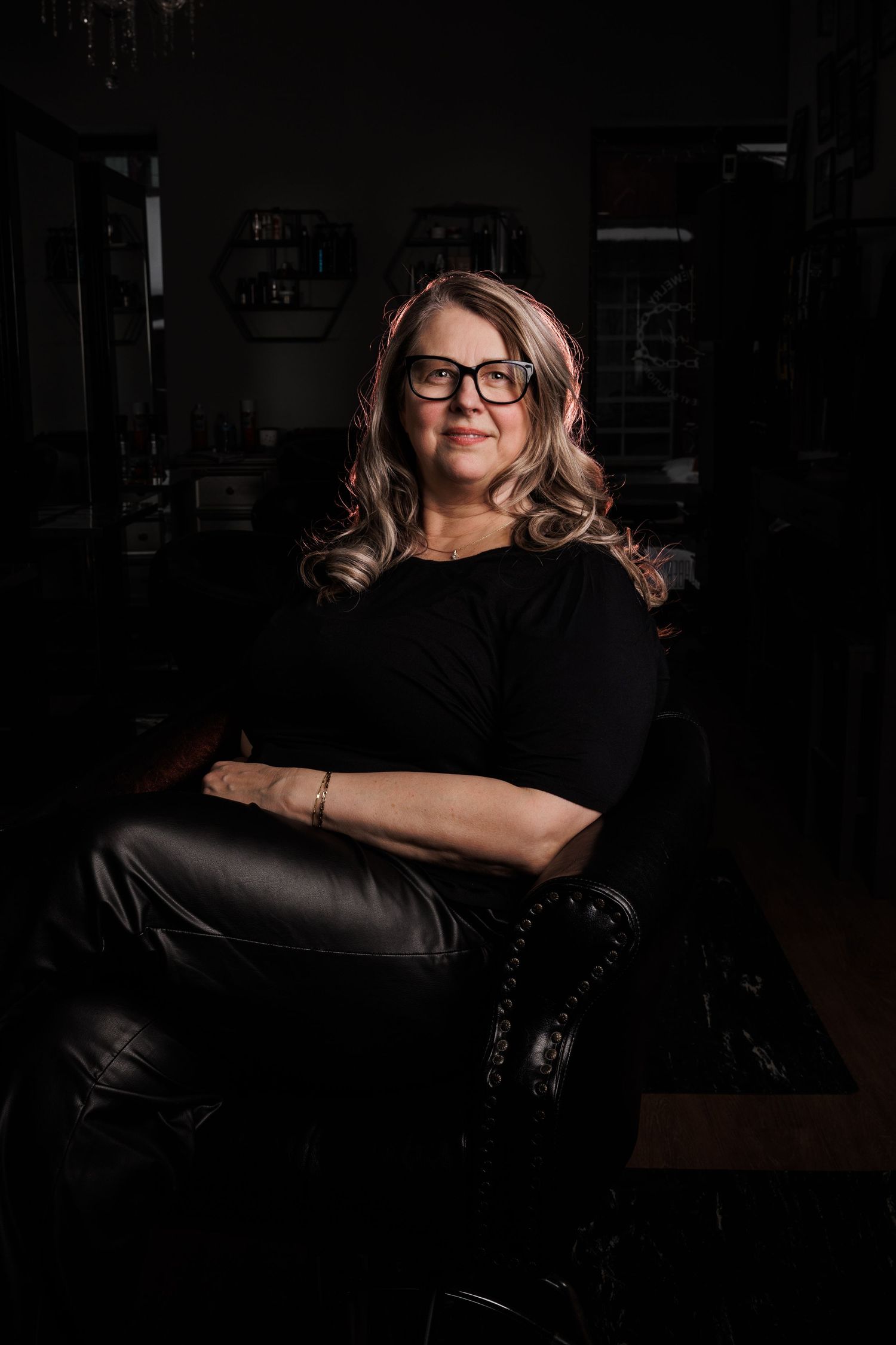 A confident woman with glasses and highlighted hair sits in a dark leather chair wearing a black outfit.