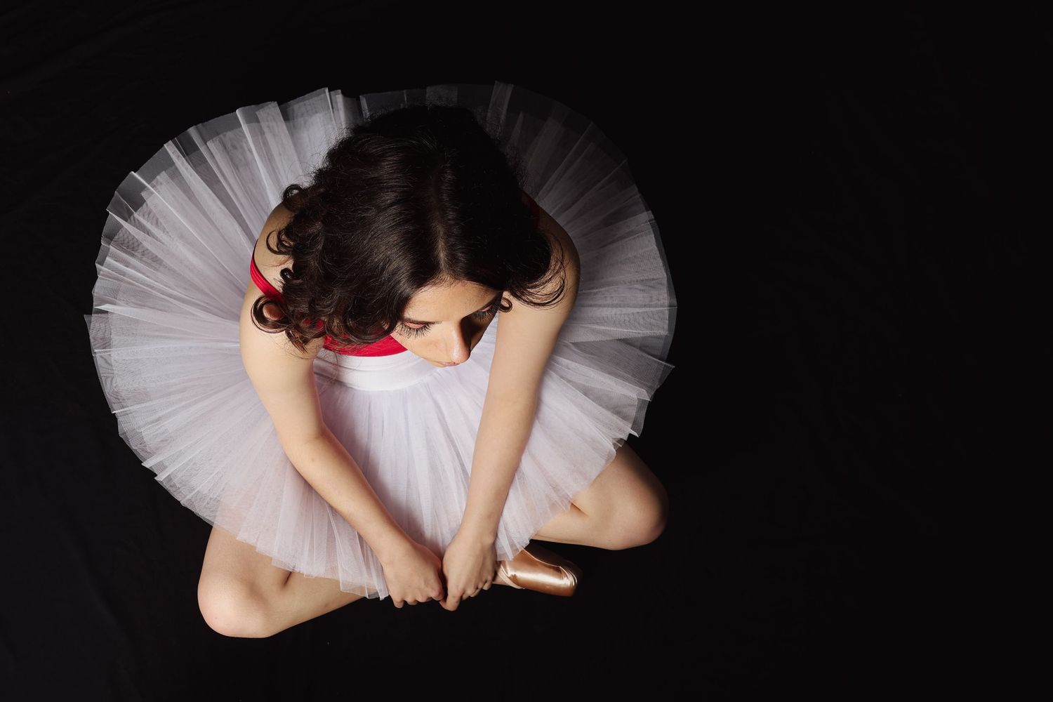 Aerial view of a ballerina in a white tutu sitting gracefully on a dark background.