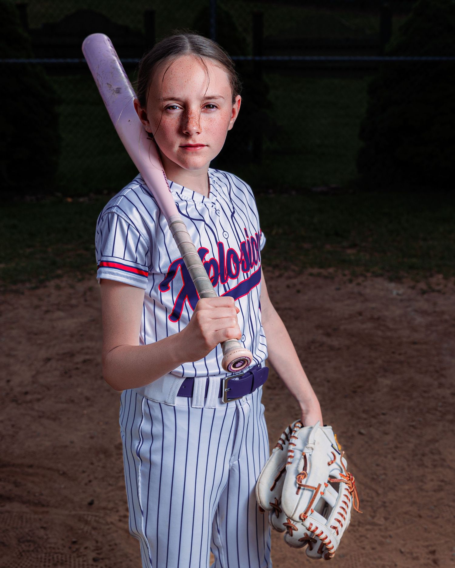 Young softball player in pinstripe uniform poses with bat and glove for sports portrait photo.