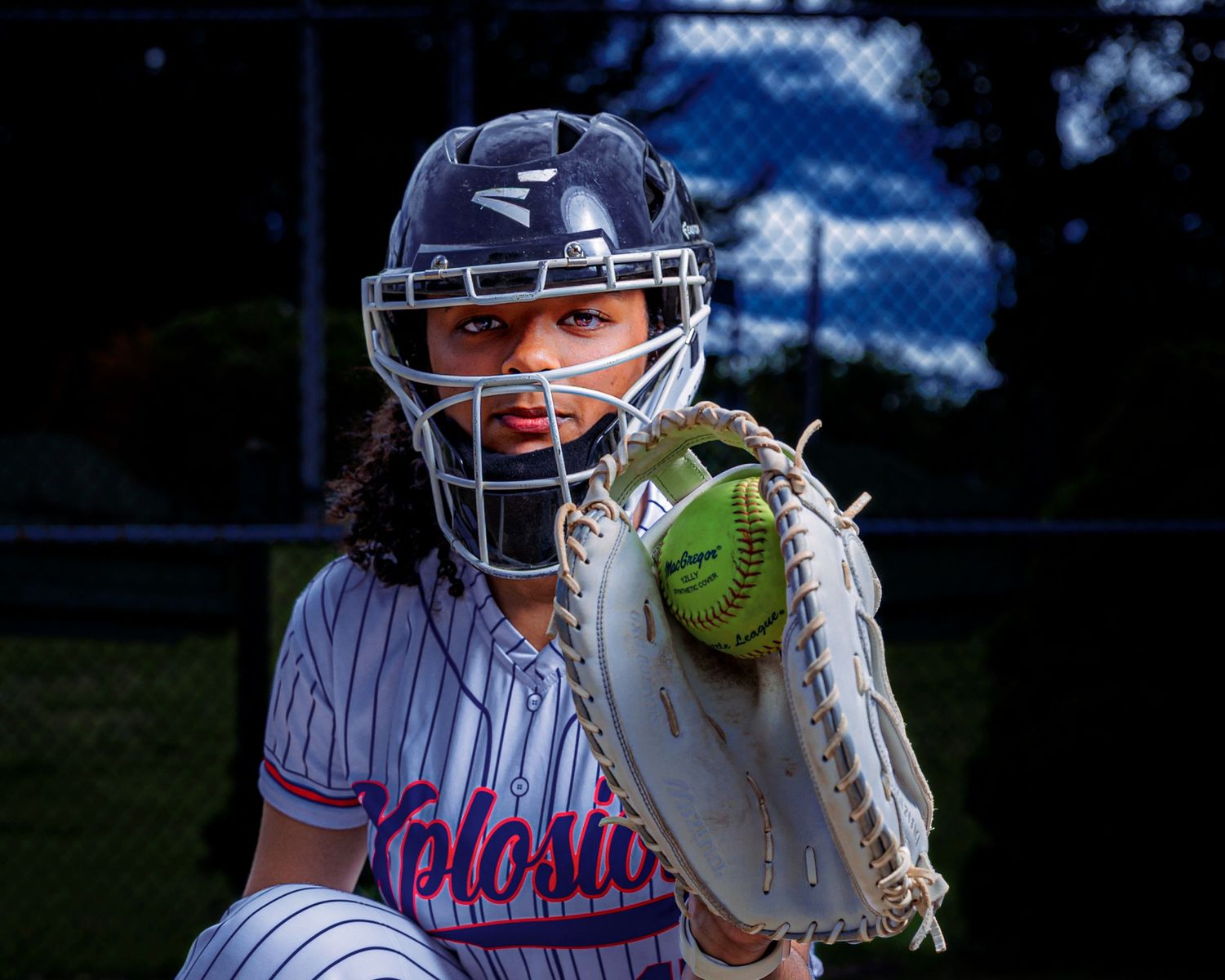 Female softball catcher in navy pinstripe uniform and helmet holds yellow ball in glove during game.