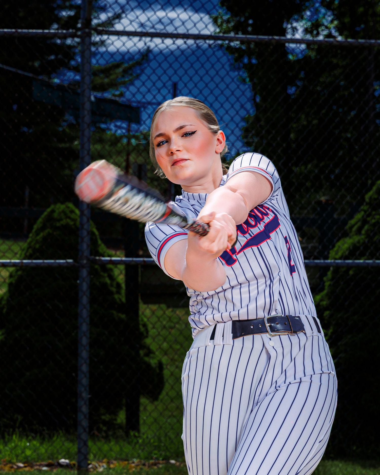 Young female softball player in pinstripe uniform swinging a bat on a sunny day near a chain-link fence.
