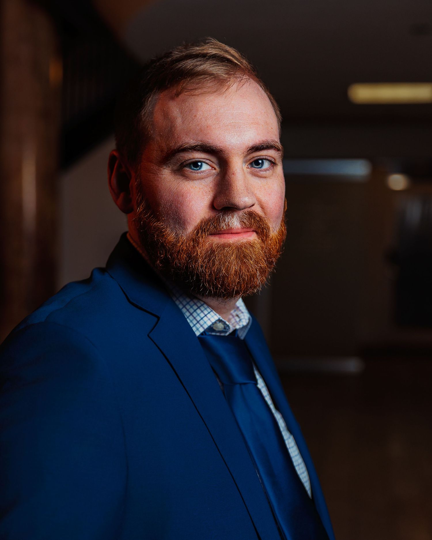 Professional man with red beard in blue suit smiling confidently in a formal portrait setting.