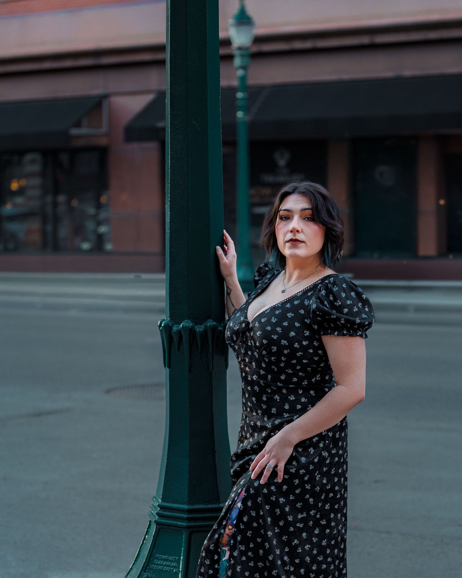 Woman in black floral midi dress poses by a green street lamp on an empty urban street at dusk.